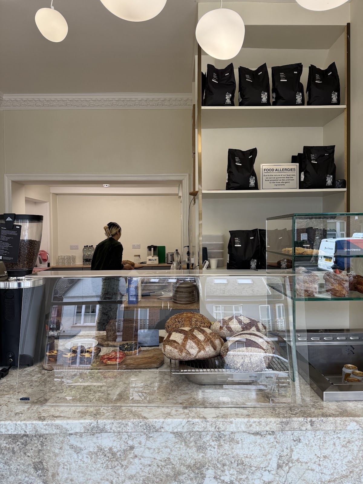 The bakery counter: sourdough loaves and pastries in a glass display case on marble, bags of coffee on shelves behind, and a member of staff working at the back bench.