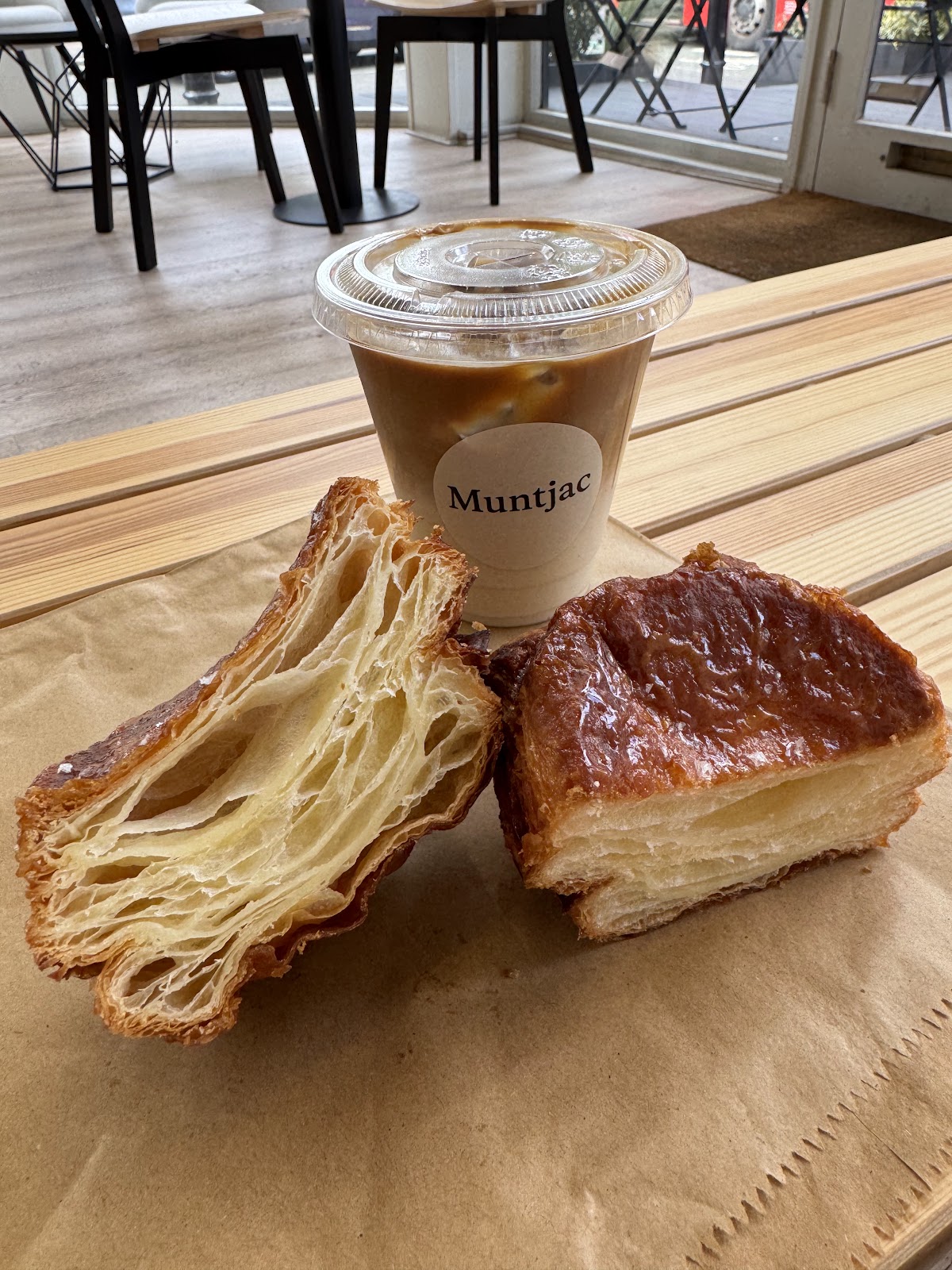 A kouign-amann broken open to show its layers, beside a whole one and an iced coffee in a cup labelled Muntjac, on brown paper on the wooden bench by the window.