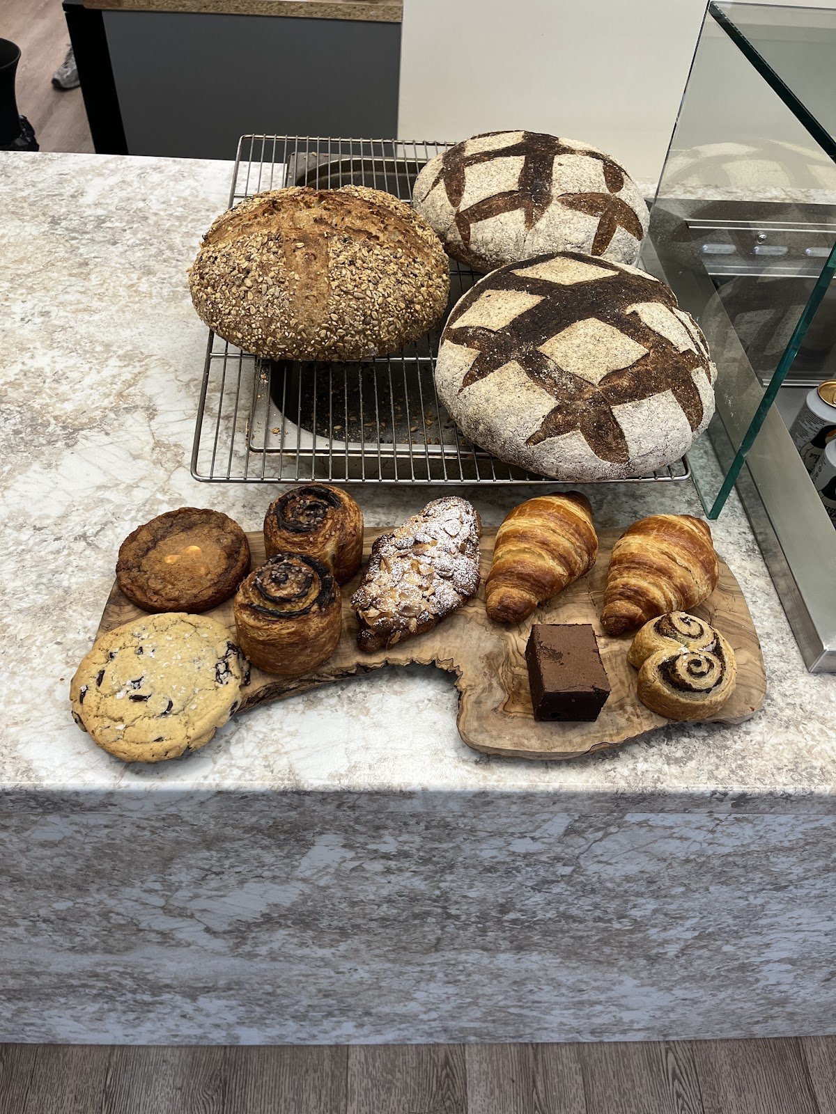 On the marble counter: two scored sourdough loaves and a seeded loaf on a cooling rack, with croissants, an almond croissant, chocolate and cinnamon swirl buns, a cookie and a brownie laid out on a wooden board.